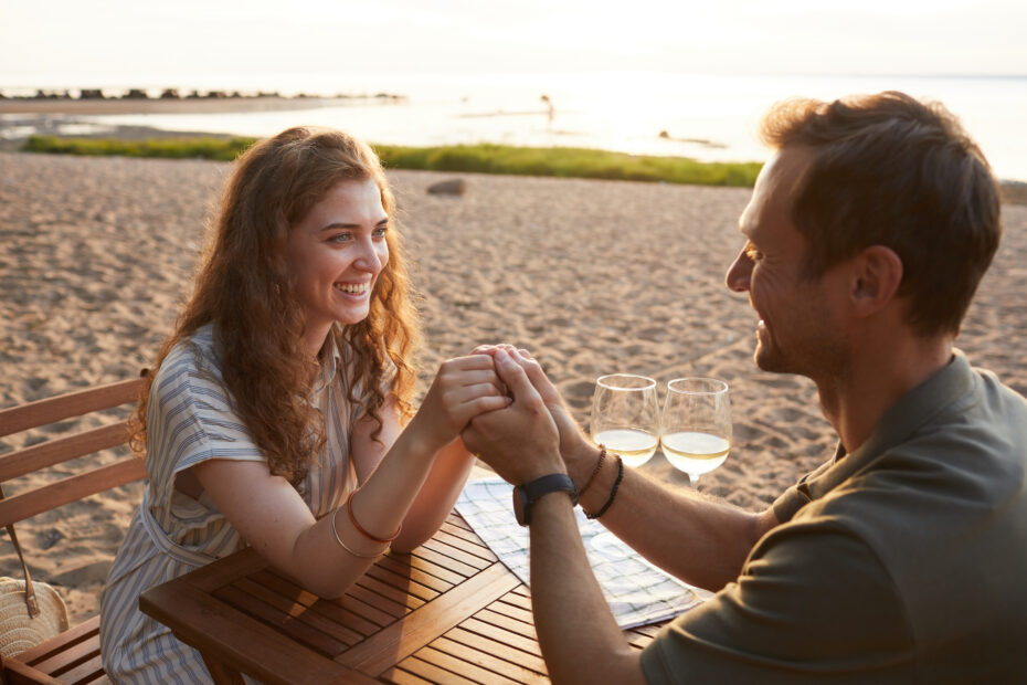 A photo of a man and woman in love sitting at a table with wine on a beach holding hands